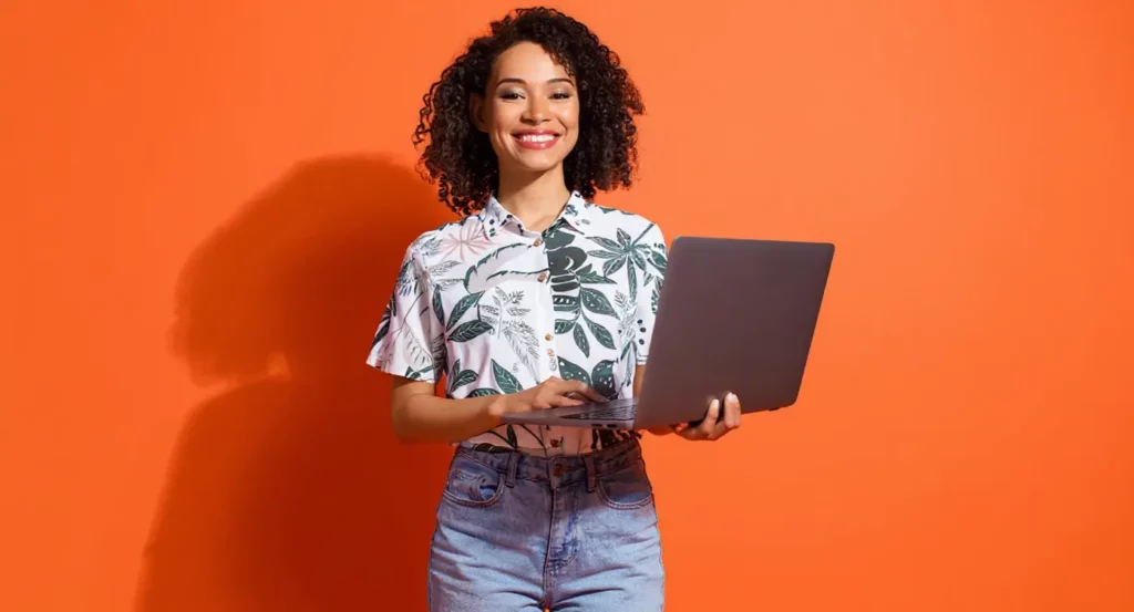 Femme souriante avec cheveux bouclés tient un ordinateur portable devant un fond orange vif.