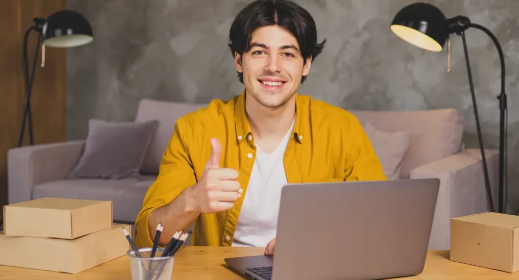 Homme souriant en chemise jaune faisant un pouce levé devant un ordinateur portable dans un bureau moderne.