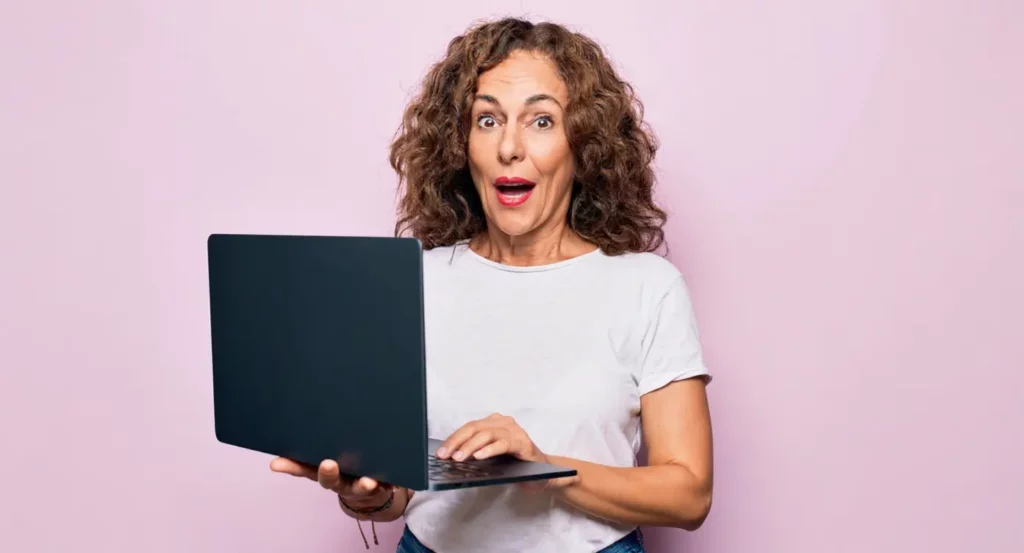 Femme souriante avec des cheveux bouclés tenant un ordinateur portable devant un fond rose.