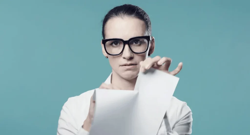 Femme en blouse blanche déchirant des feuilles de papier, avec un regard intense et des lunettes noires, sur fond bleu.