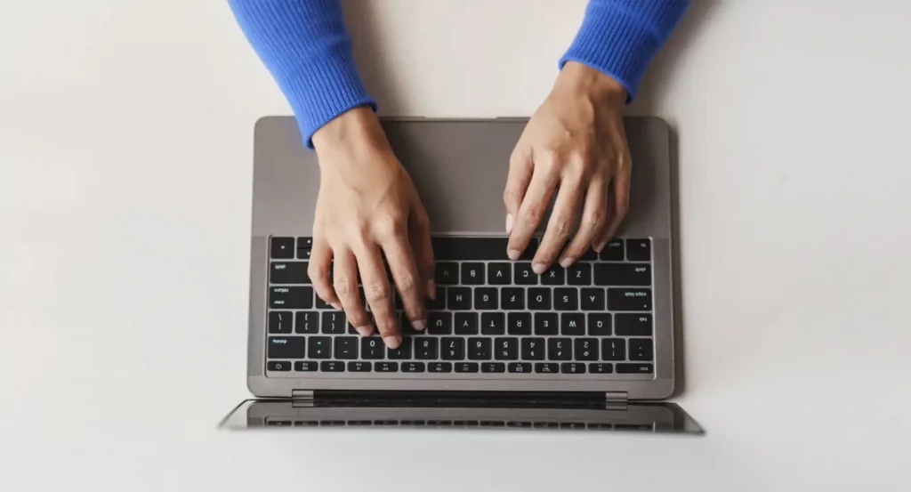 Femmme tapant sur un clavier d'ordinateur portable, portant un pull bleu, sur une table blanche.