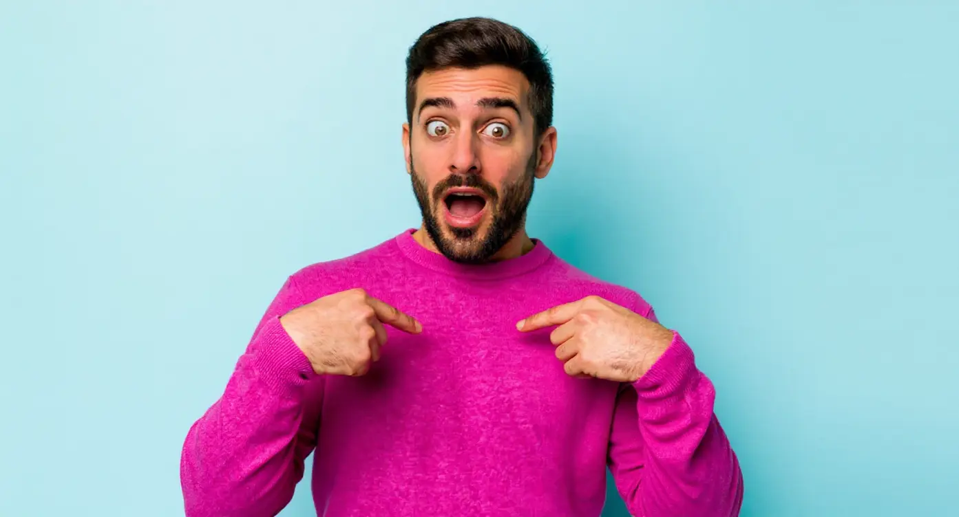 Homme souriant avec un pull rose vif, pointant vers sa poitrine sur fond bleu.