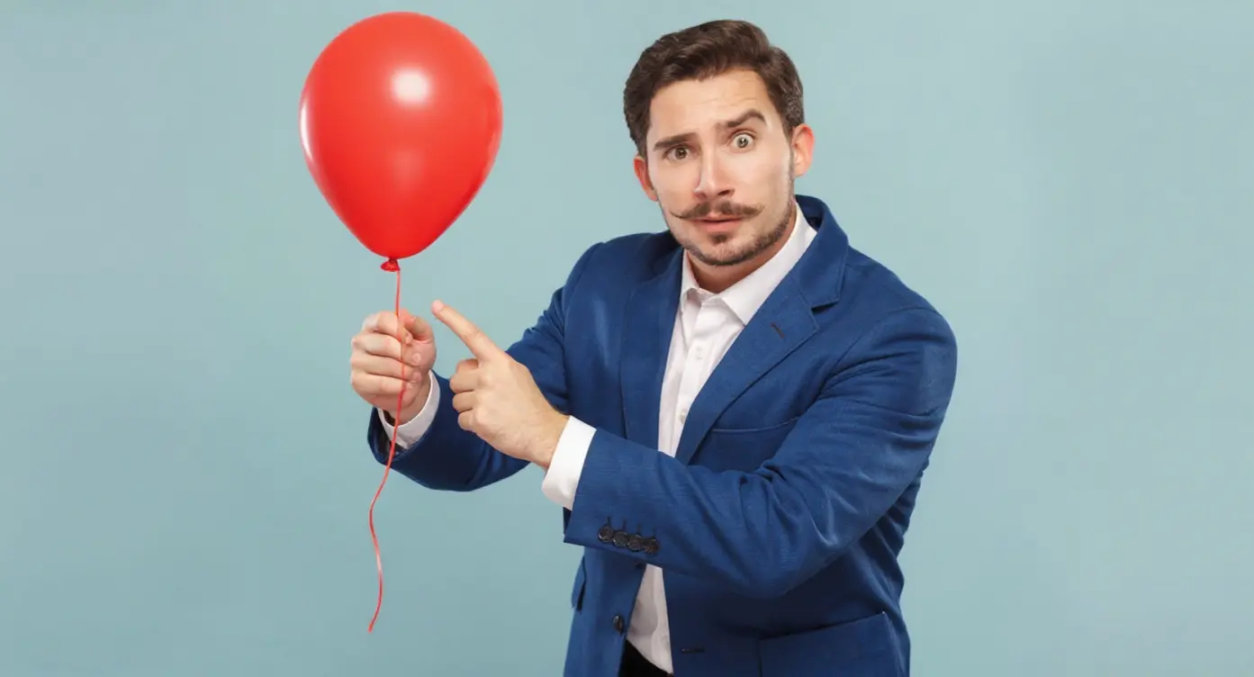 Homme en costume bleu tenant un ballon rouge, montrant une expression surprise et pointant du doigt.