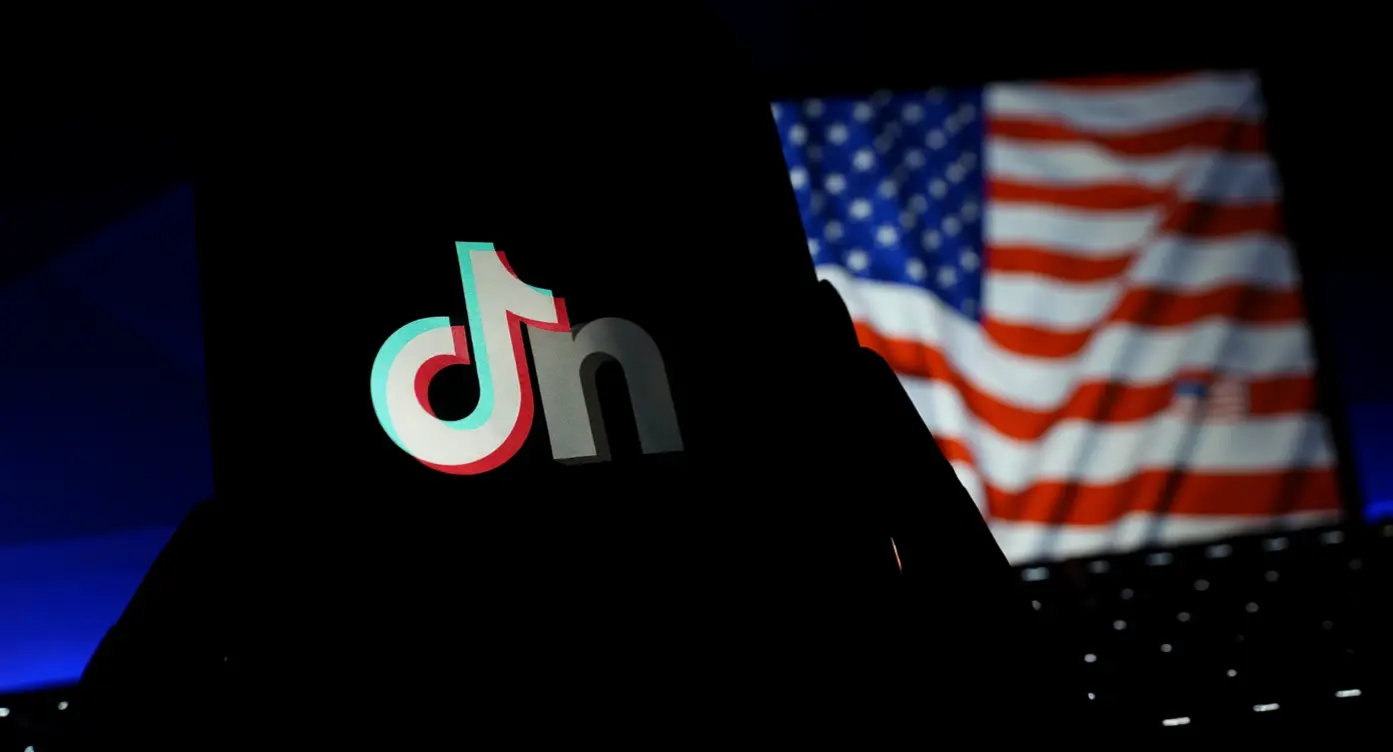 Logo de TikTok en silhouette avec un drapeau américain en arrière-plan flou, symbolisant la culture numérique et les réseaux sociaux.