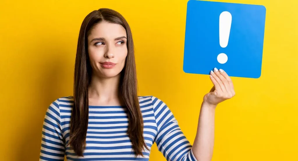 Femme souriante devant un fond jaune, tenant une pancarte bleue avec un point d'exclamation.