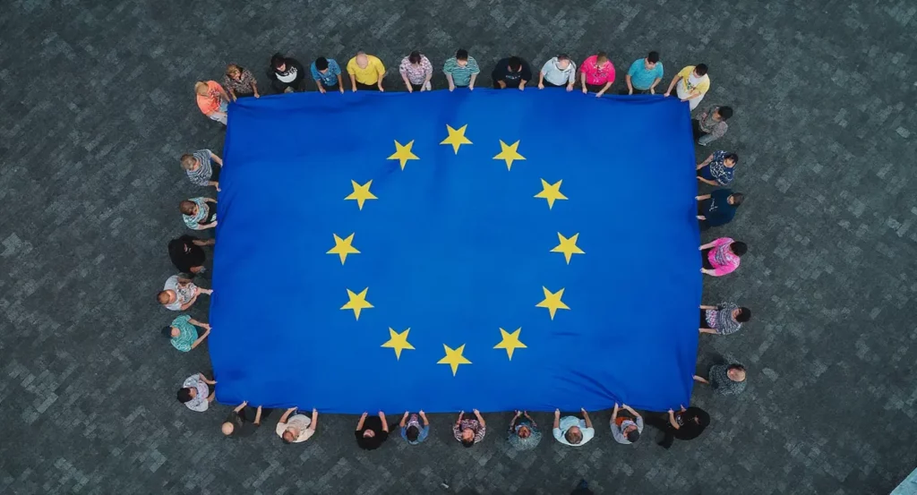 Des personnes tiennent un grand drapeau bleu de l'Union européenne avec des étoiles jaunes sur un sol en pavés.