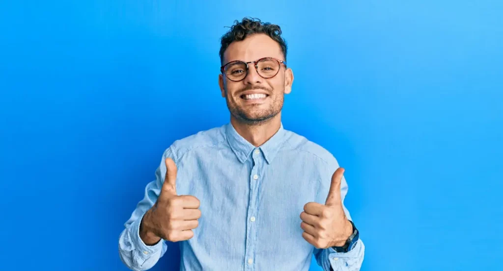 Homme souriant en chemise bleue, levant les pouces sur fond bleu, exprimant la satisfaction et la positivité.