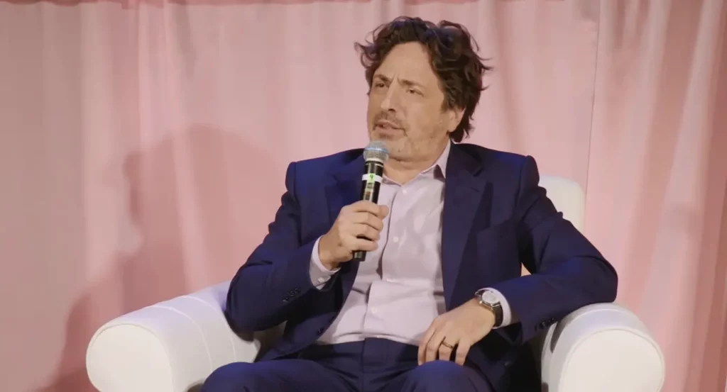 Intervenant en costume, s'exprimant avec un micro sur scène devant un fond rose.