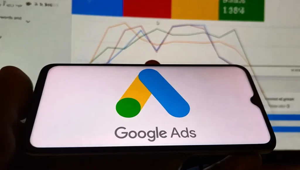 Logo de Google Ads affiché sur un smartphone avec des graphiques analytiques en arrière-plan.