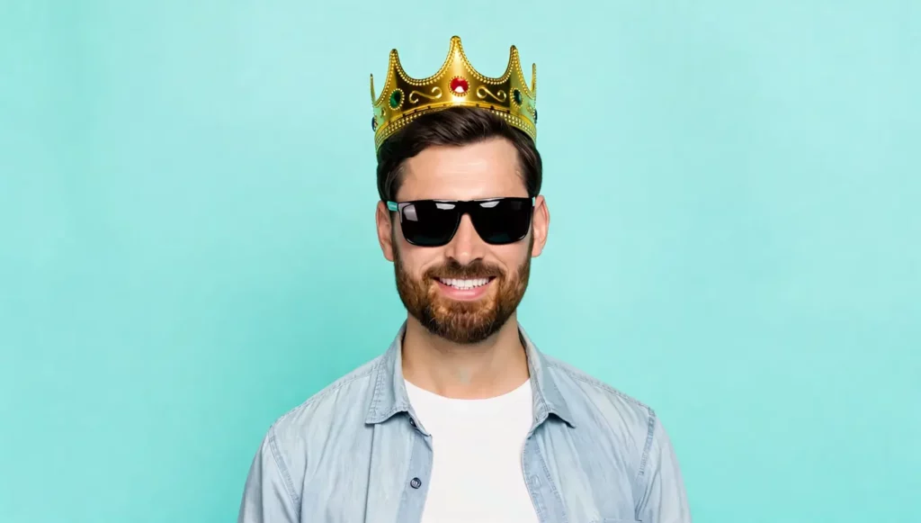 Homme souriant avec une couronne dorée et des lunettes de soleil sur fond turquoise.