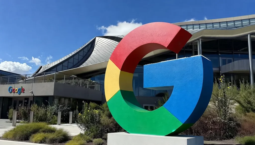 Logo coloré de Google devant un bâtiment moderne sous un ciel bleu avec quelques nuages.